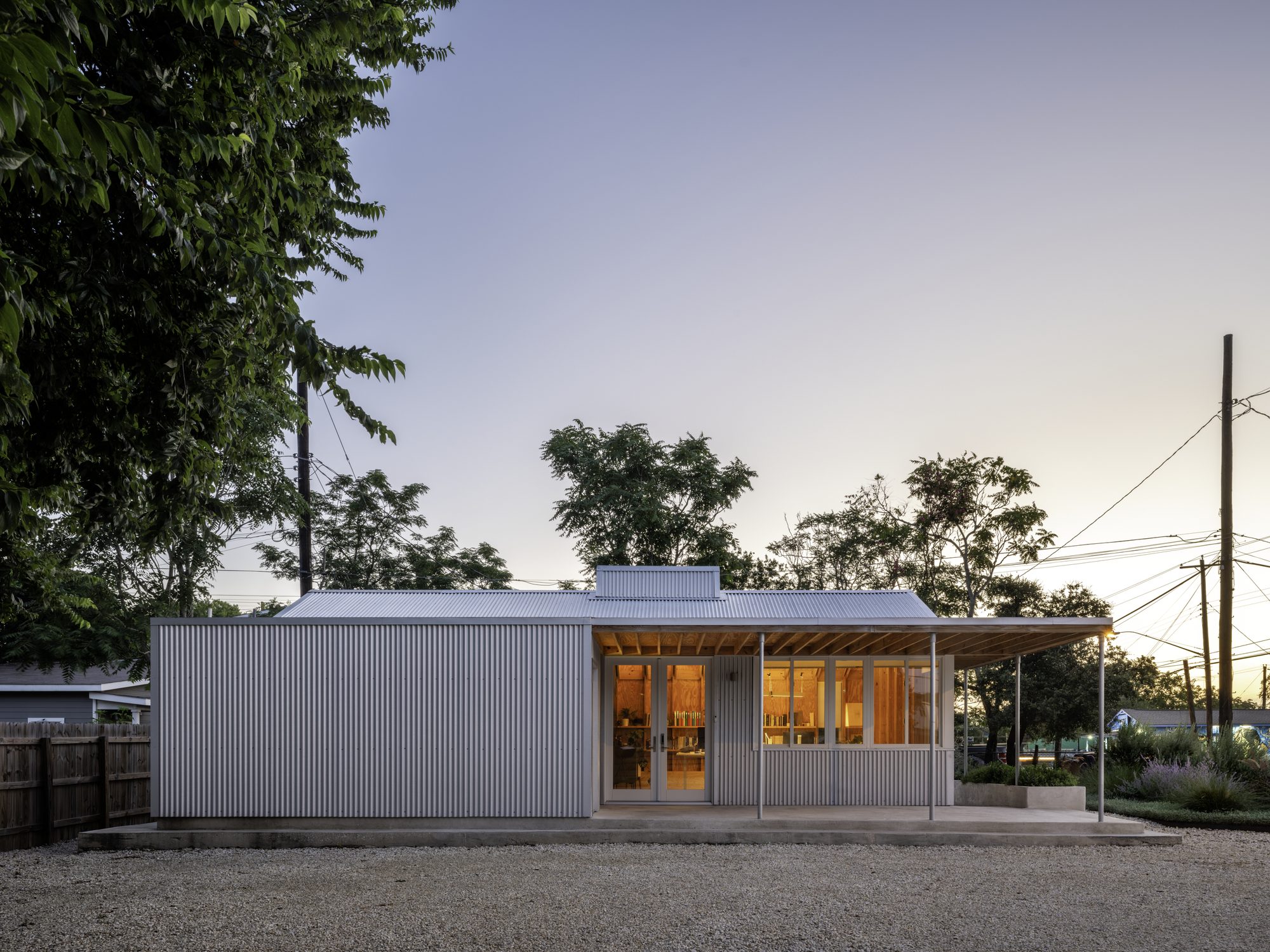 Exterior of the Architect's Studio at dusk — a small corrugated metal building with a deep porch and warm interior light visible through full-height windows, set on a gravel court framed by mature trees and overhead utility lines