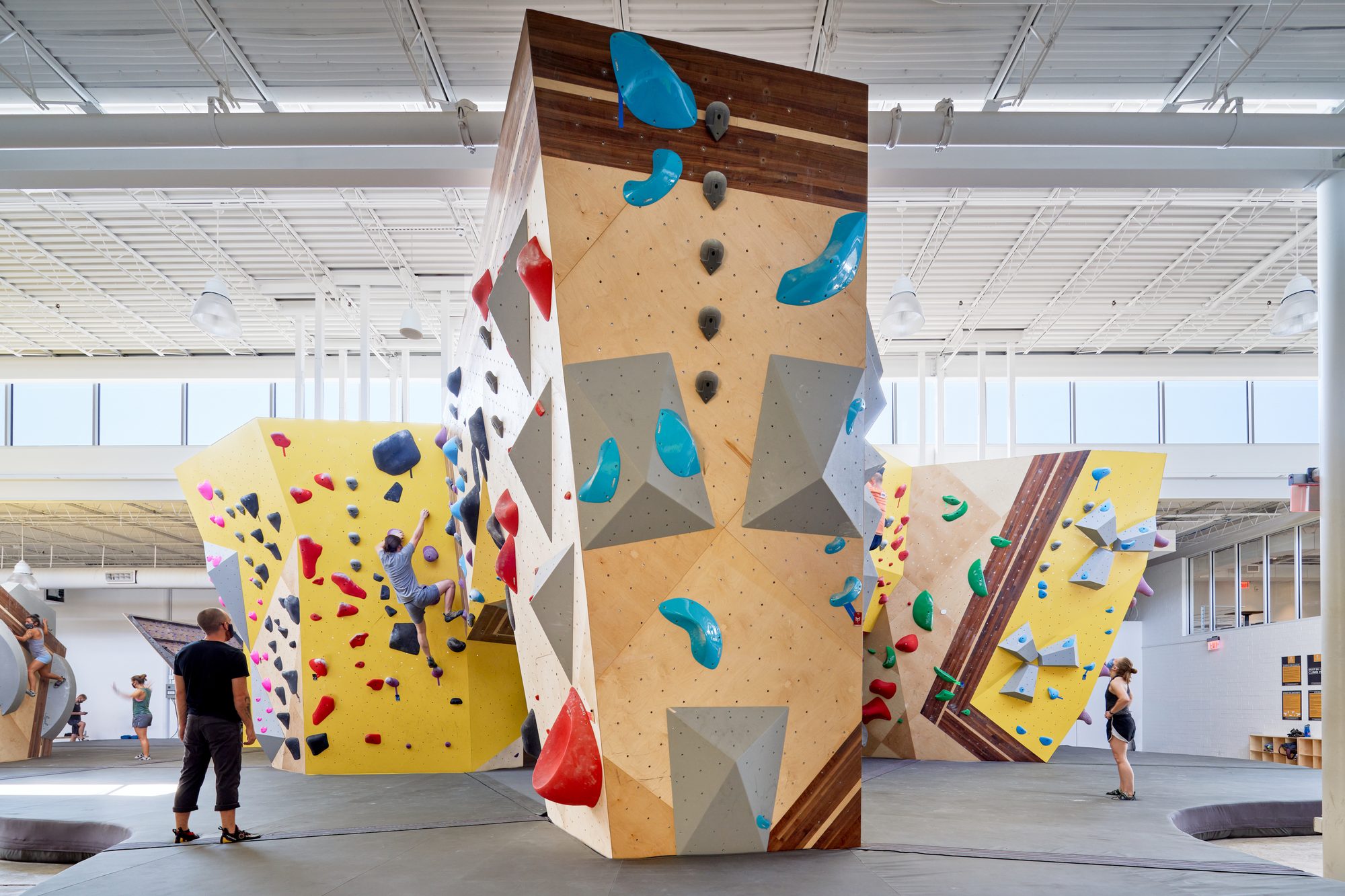 Tall central faceted boulder at Crux Central with bright teal and orange holds, a climber on the wall mid-route, and two figures standing at the base — the wider gym and faceted boulders visible to either side
