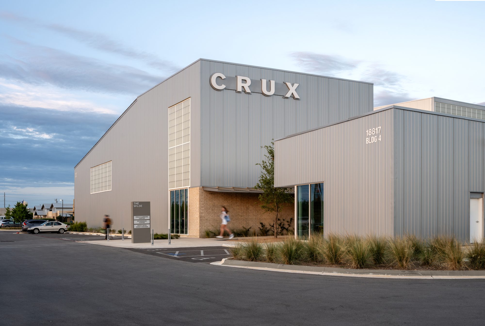 Exterior corner of Crux Pflugerville climbing gym at dusk, showing the metal-clad facade, the illuminated CRUX wordmark above the entry, and a brick base anchoring the entry
