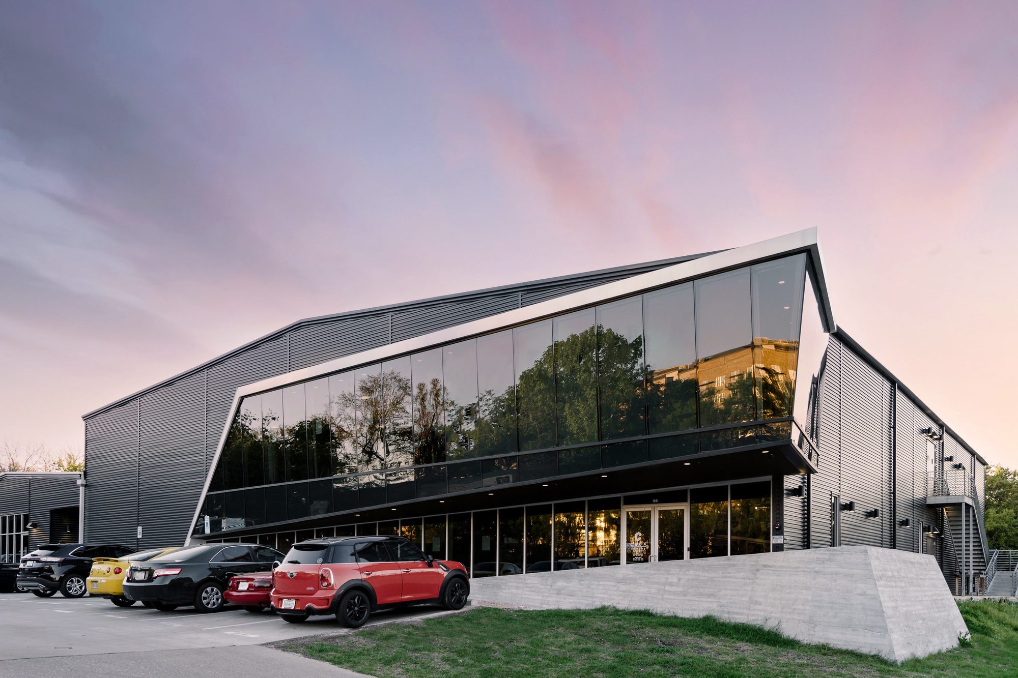 Dusk exterior of Crux South Congress climbing center showing the cantilevered glass volume jutting toward the parking lot, dark corrugated metal cladding wrapping the existing shell behind, and a stepped concrete plinth at the entry under a pink-and-purple sunset sky