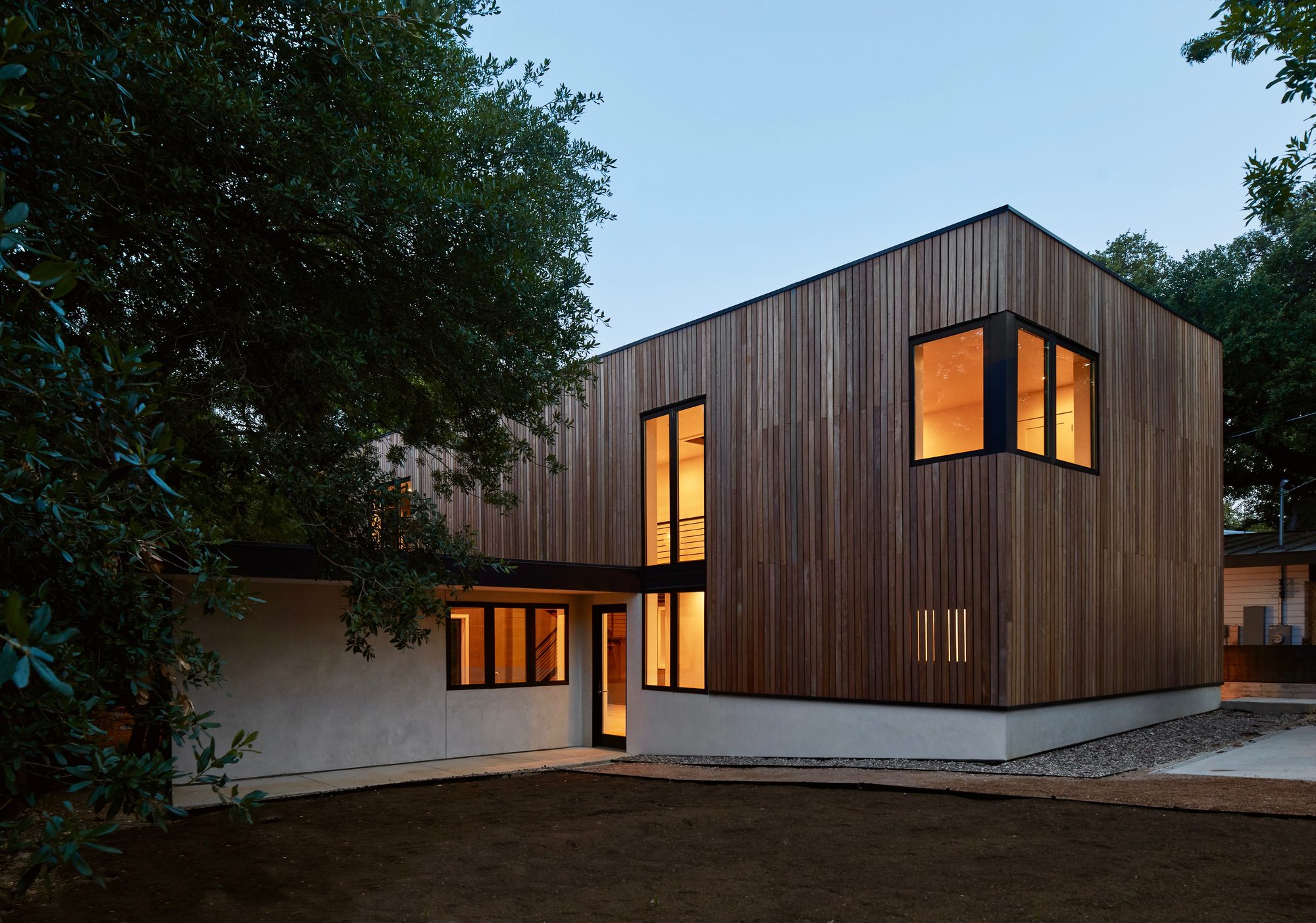 Twilight exterior of the Jesse Street House showing the cedar-clad upper volume glowing against the deep blue sky, the stucco-and-block ground floor below with warm interior light visible through black-framed windows, and a large oak tree in the foreground