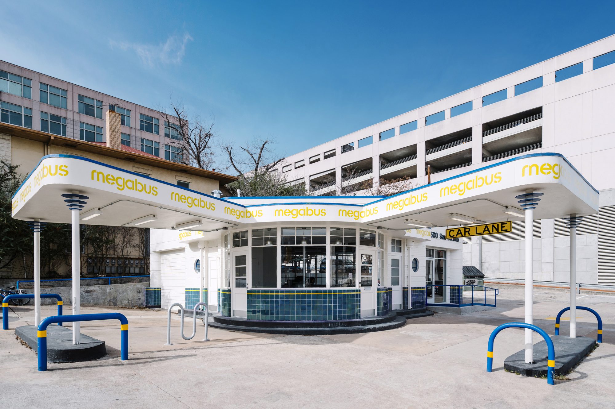 Wide exterior view of the Megabus Station from across the lot, showing the restored 1942 Streamline Moderne canopy with megabus wordmark lettering, the curved entry pavilion below with green tile band, blue-and-yellow bollards, and a downtown parking garage rising in the background